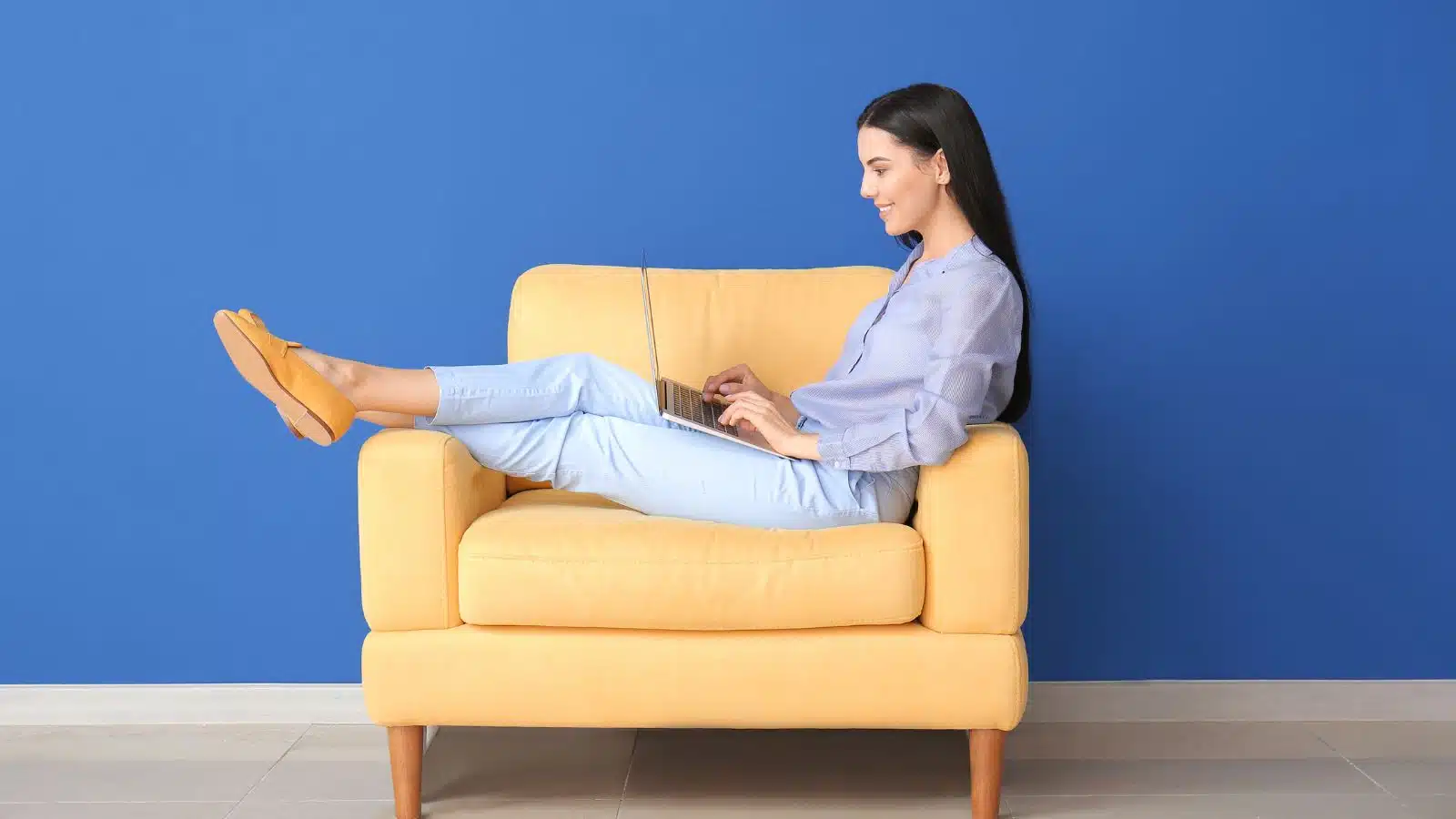 young woman sitting on a yellow chair on her laptop smiling