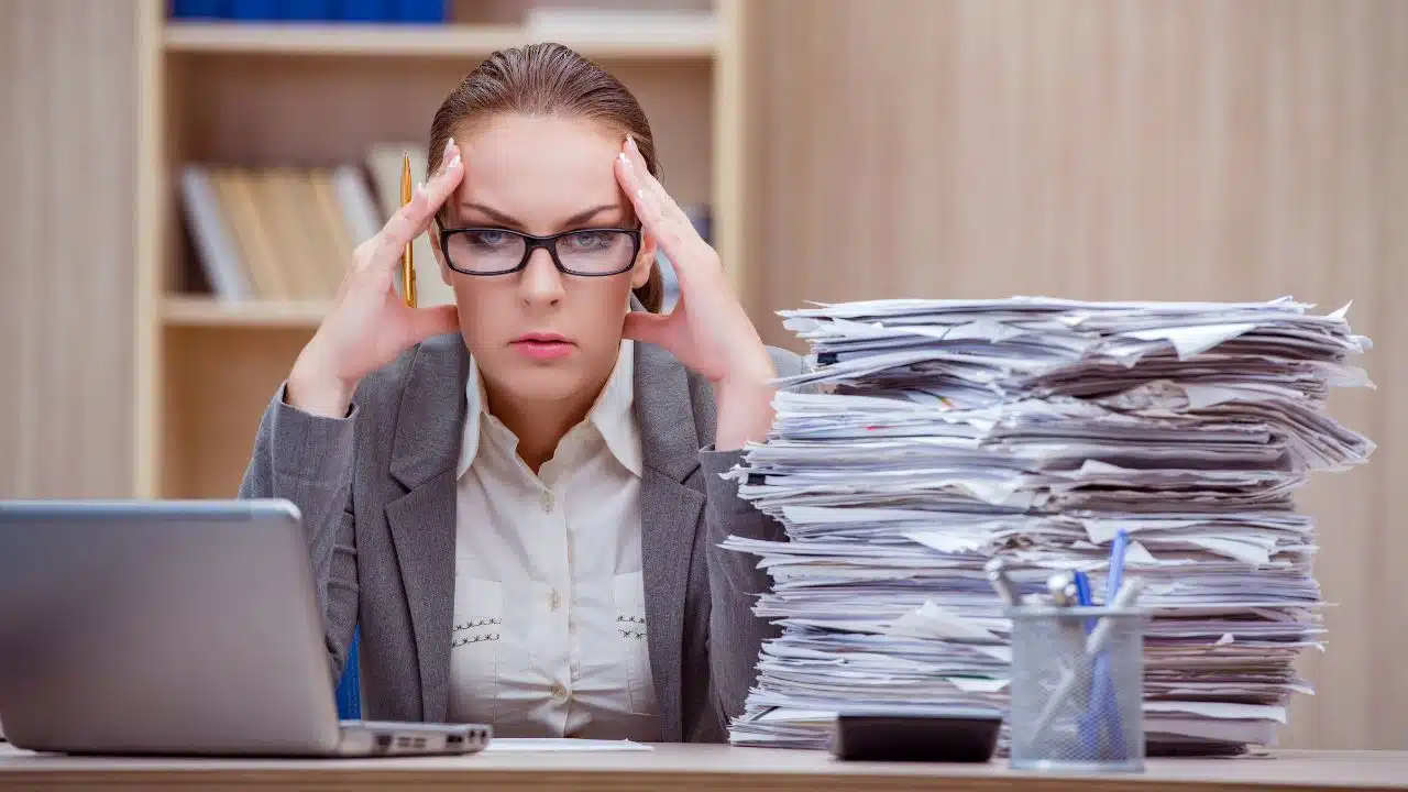 beyond stressed woman at her desk in an office with a giant stake of paperwork