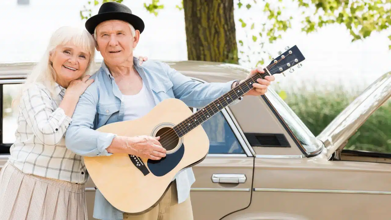 happy retired couple with guitar standing next to a car outdoors