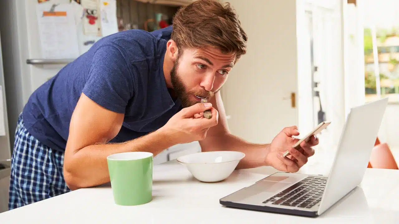 man eating breakfast staring at phone and computer