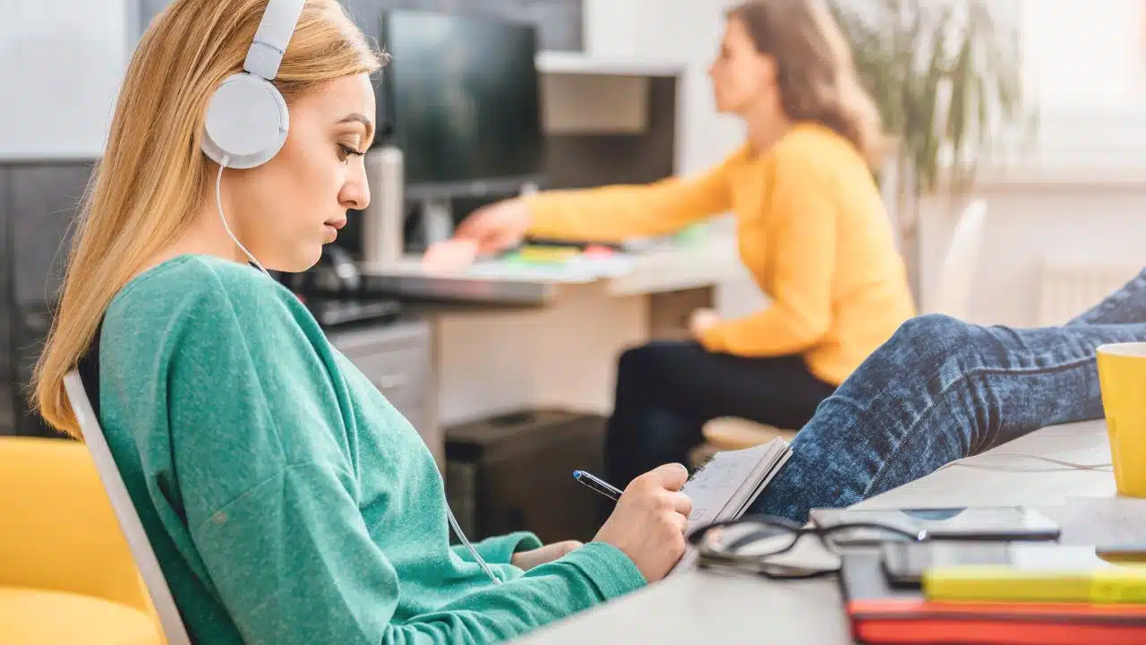 woman with feet up on desk writing in notebook with headphones